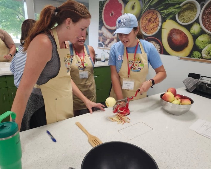 Two woman, both wearing aprons, standing along a worktable using an apple peeler. Another woman is behind them, watching them.