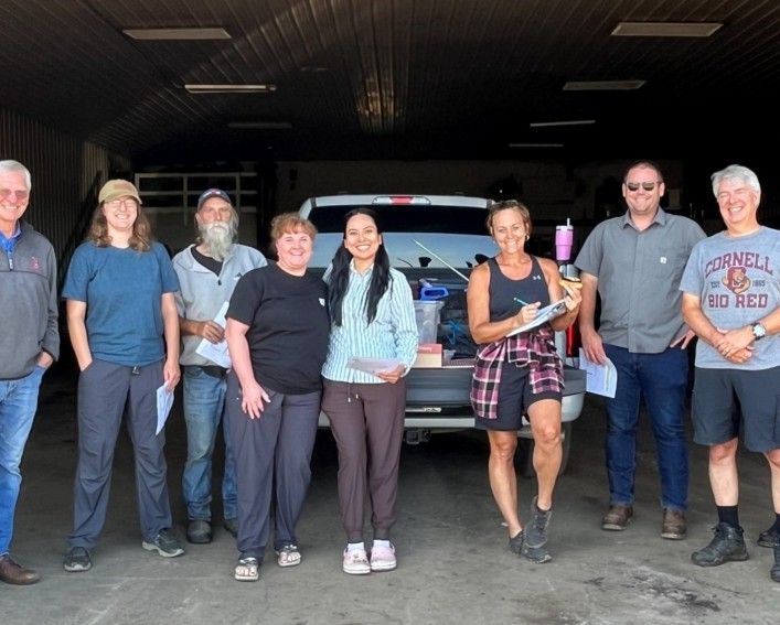 Nine smiling people standing next to each other at the tailgate of a pickup truck parked within a barn. The woman in the middle is holding a clipboard.