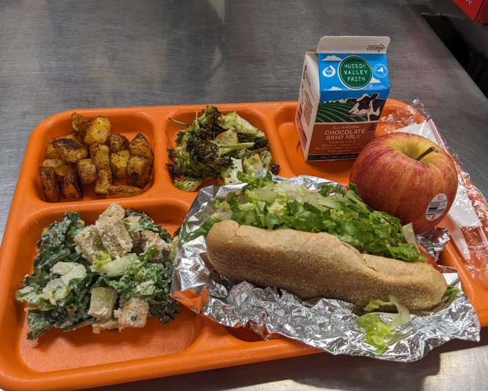 Orange plastic multi-compartment lunch tray with an apple, chocolate milk carton, roasted broccoli and potatoes, caesar salad, and a sub sandwich topped with fresh chopped lettuce.