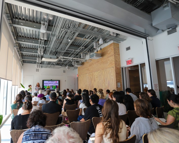 A group of around 75 individuals gather in seated rows facing a screen with two people presenting.