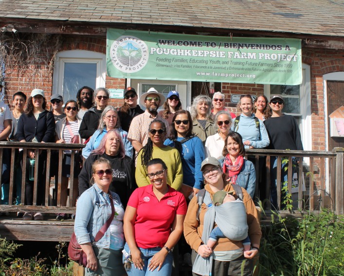 A group shot of nearly two dozen people outside in front of a brick building with a banner hanging from it that says "Welcome to Poughkeepsie Farm Project" in English and Spanish.