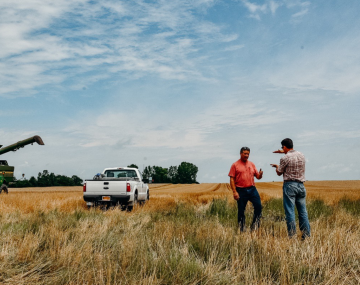 two men stand in a field with a green combine behind them