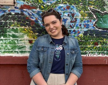 A woman stands smiling in front of a brightly painted mural. She’s wearing a denim jacket over a navy shirt and a light-colored skirt. Her hands are clasped in front of her, and sunglasses rest on her head. The mural behind her features colorful abstract shapes, leaves, and flowers in blues, greens, reds, and yellows.