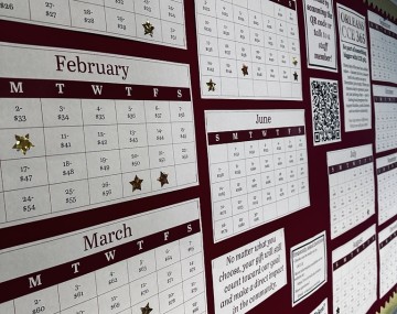 A photo of the Orleans CCE 365 donor wall display. Several printed monthly calendars are arranged across a red bulletin board with gold trim. Some dates on the calendars have gold star stickers indicating donated days. A section of the display includes a QR code for donating and informational text about the fundraiser. An exit sign and part of the hallway ceiling are visible at the top.