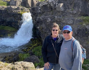 Two adults standing together on a rocky overlook in front of a small waterfall cascading between rugged cliffs. The woman is wearing sunglasses, a black jacket, and jeans. The man is wearing sunglasses, a blue baseball cap, a gray sweatshirt, jeans, and a backpack. Moss-covered rocks and flowing water are visible below, with steep rocky hills rising in the background under a clear blue sky.