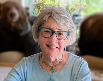 An older woman with short gray hair and teal glasses smiles at the camera while seated indoors. She is wearing a light blue long sleeve shirt and layered turquoise jewelry. The background is softly blurred, showing large nature themed artwork and two bears sitting behind her.