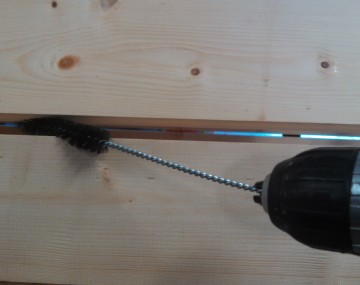 Close up of a drill with a brush chucked in. The thin brush fits between two wooden slats in a root barrel washer.